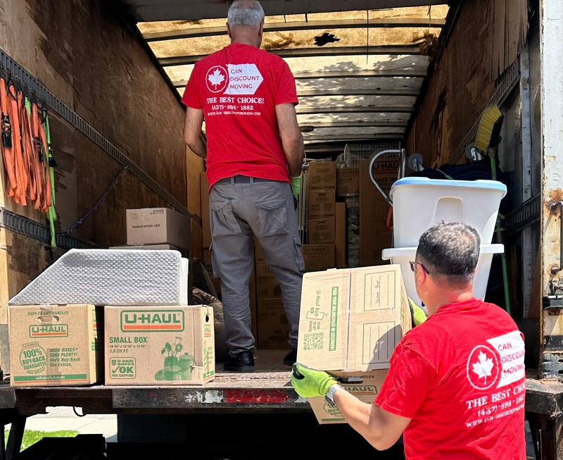 Professional mover lifting packed moving boxes into a moving truck while preparing a family home for relocation. (116 chars)